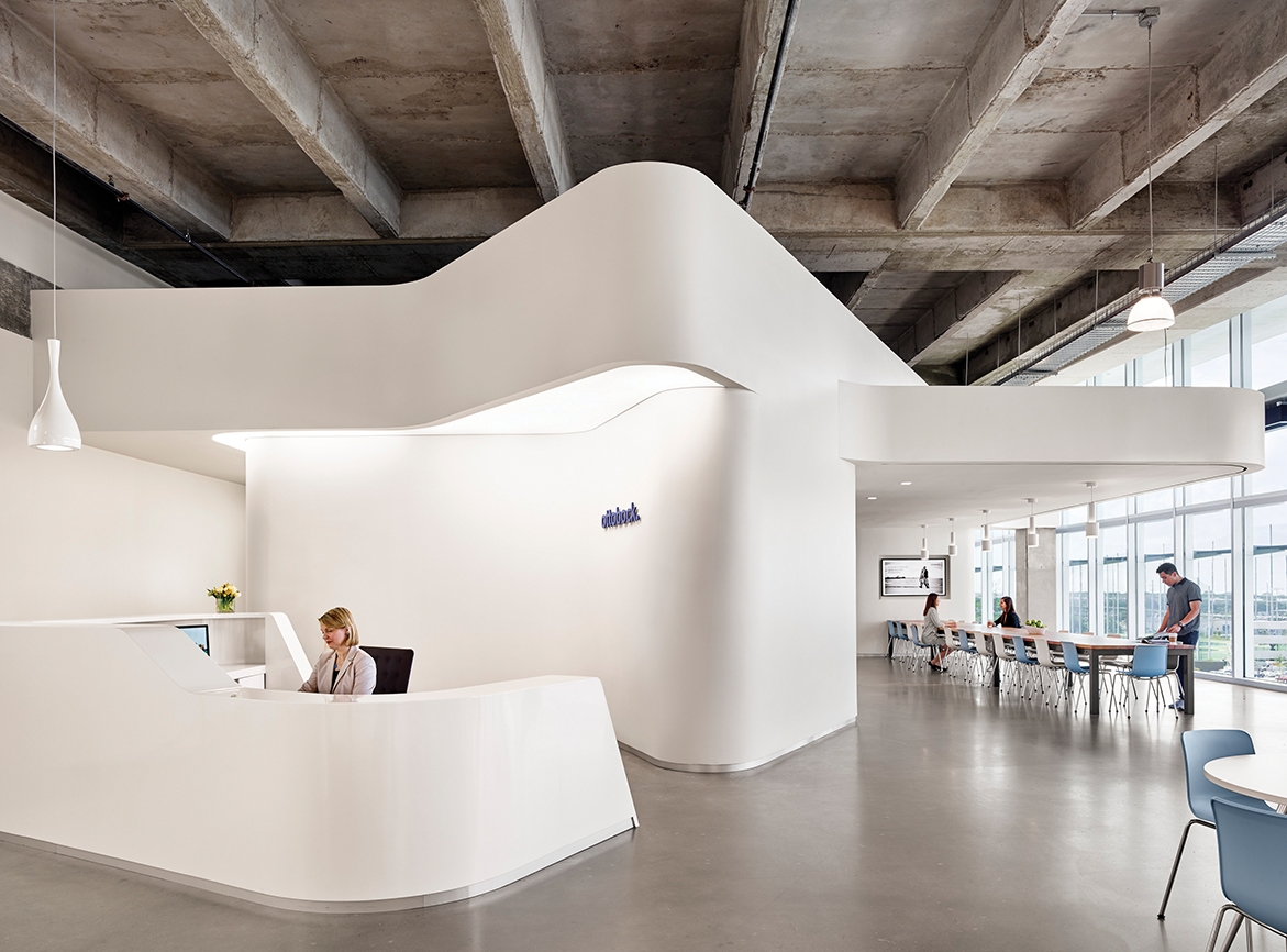 Curved white reception desk welcoming visitors into a bright space with ample daylight. Cream and light blue chairs line a long table in the background.