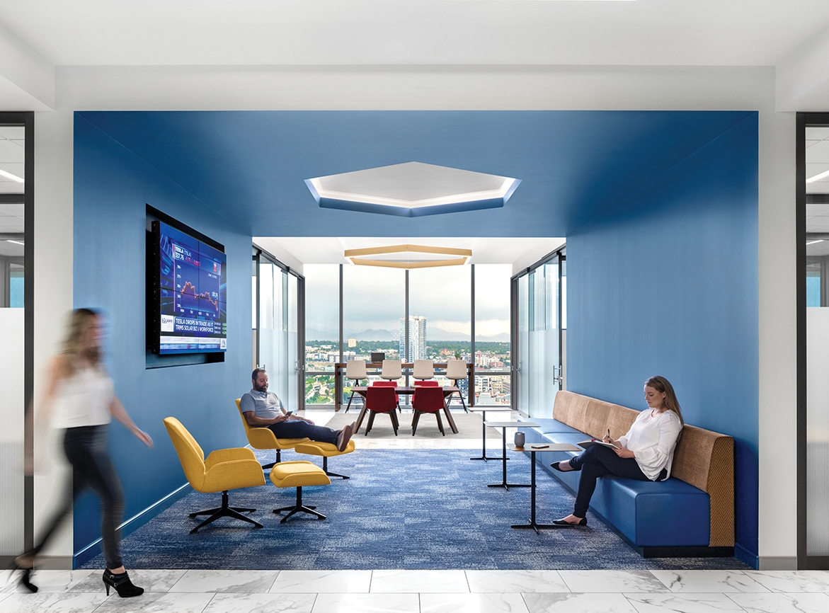 View into a blue themed breakout space with blue carpet, ceiling, and walls. A blue vinyl and wood bench is along the right wall with some small tables. Yellow chairs with footrests are positioned along the left wall. In the background are more tables and chairs.